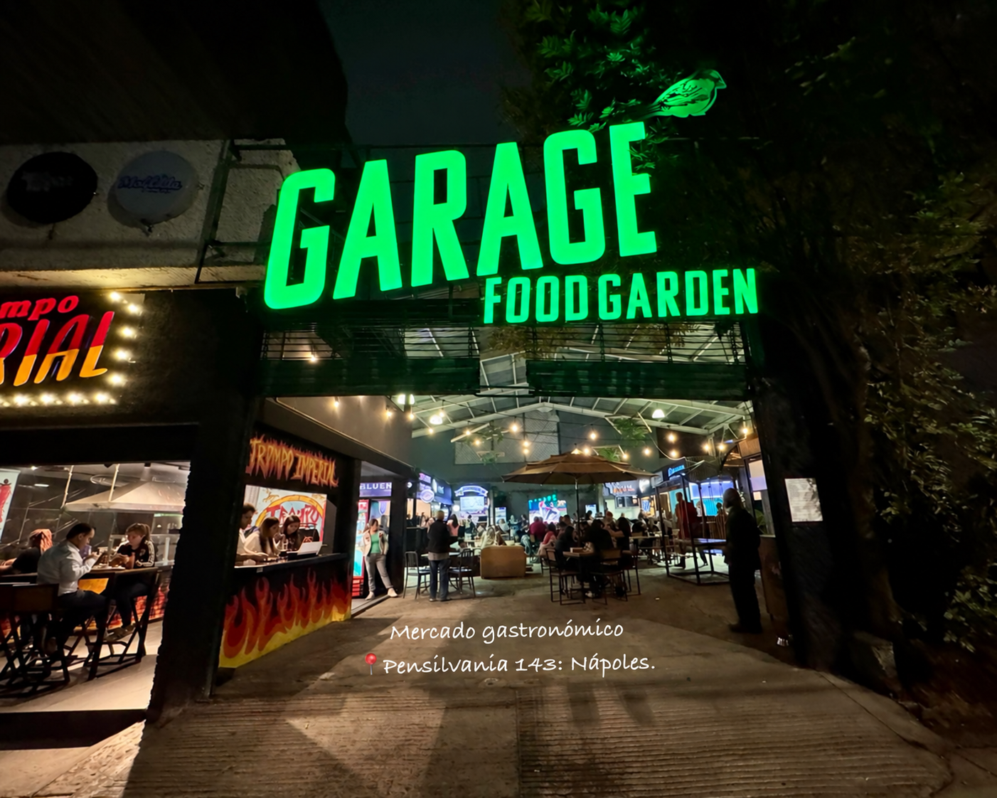 Entrada de Garage Food Garden de noche, con el letrero verde y mesas iluminadas